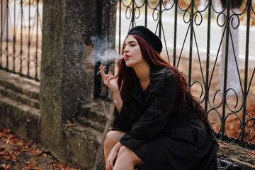 A girl in retro coat smoking cigarette sitting on fence in the autumn park