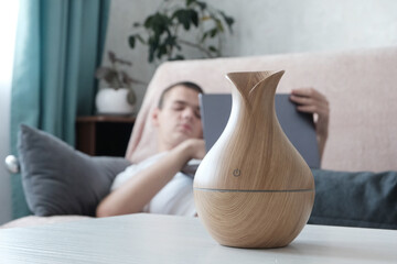 Wooden bamboo electric diffuser on a light table. The concept of humidifying and aromatizing the air in the house. Young man sleeping in the background in blur background. - obrazy, fototapety, plakaty