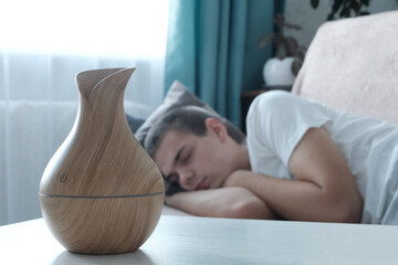 Wooden bamboo electric diffuser on a light table. The concept of humidifying and aromatizing the air in the house. Young man sleeping in the background in blur background.