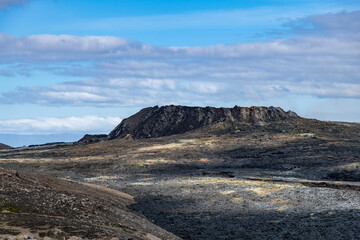 Fototapeta na wymiar Fissure vent in Geldingadalir volcano area that was active in 2021 near mountain Fagradalsfjall, Iceland with black lava field in forefront against a white clouded blue sky