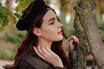 A girl with earrings in retro beret posing near tree in the autumn park, french style