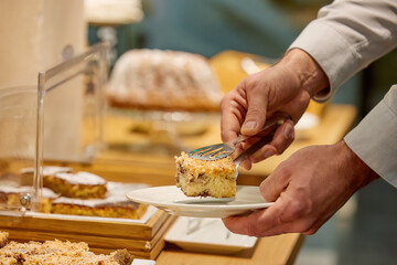 Man in white shirt at breakfast in hotel, scooping up cake among other food, sweet breakfast. Hand in white shirt, blurred background. 