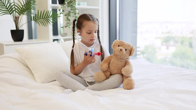 Smiling girl using toy syringe for injecting fluffy teddy bear with plastic stethoscope in ears on comfort white bed. Beautiful caucasian child imagining herself as doctor and performing procedures.