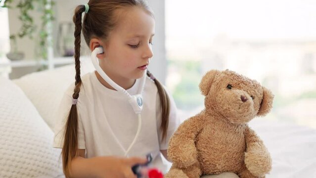 Small child pretending to be nurse and giving injection to brown teddy bear with toy needle. Charming caucasian girl in white bed examining patient. Concept of treatment and creative games.
