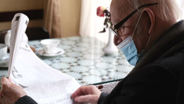 Sick old grandfather in a blue protective mask sits at the kitchen table and reads the news in the newspaper.