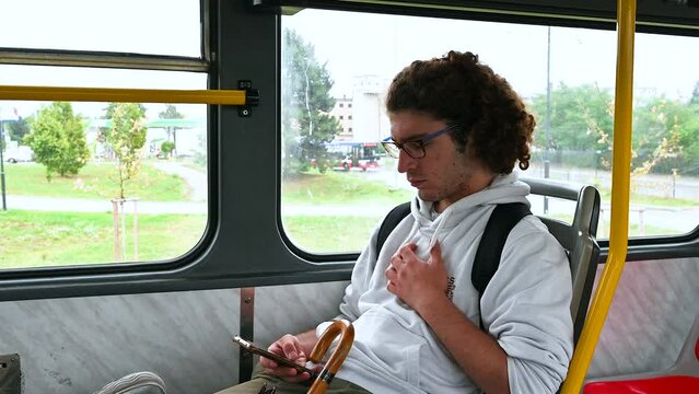 A young Caucasian man with curly hair and glasses is sitting on the bus. He is busy consulting his cell phone. Concept of use of technology by young people.