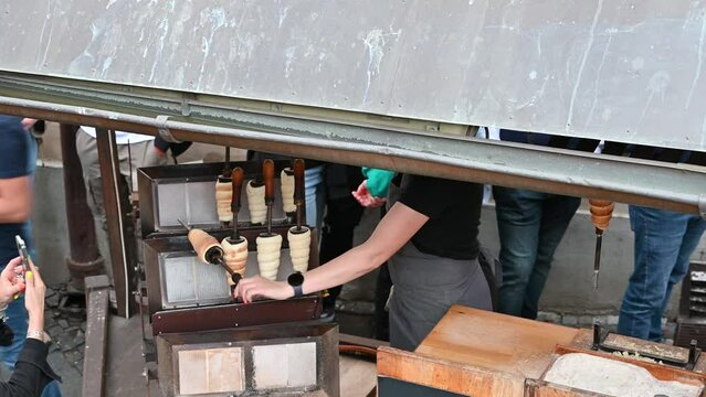 At a street food stall in Prague preparation and sale of cimney cacke, in the local language Trldo. The dough cooks while rotating on the wooden support. A tourist films him with her cell phone