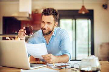 Fototapeta premium Young man going over bills and payments while drinking coffee in the kitchen