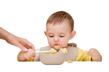 Mother teaches toddler baby boy to eat porridge with a spoon while sitting in a child chair, isolated on a white background. Mom helps the child to eat with a spoon from a plate, kid aged one year