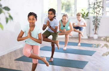 Poster Im Rahmen Children practicing yoga, standing in Tree Pose, asana Vriksasana, leg in half lotus position, hands in Namaste. © JackF