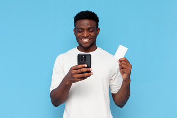 african american guy in white t-shirt uses smartphone and holds credit card on blue isolated background - obrazy, fototapety, plakaty