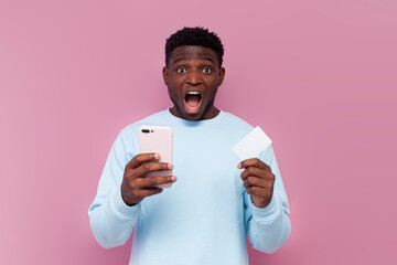 shocked african american man in blue sweater uses smartphone and credit card on pink isolated...