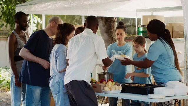 At homeless shelter team of voluntary people dedicatedly packaging food parcels to give to less fortunate. Poor and needy individuals patiently waiting to be served free meal by charity workers.