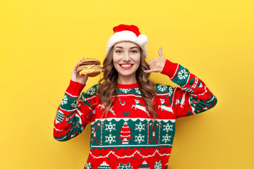 young cute girl in christmas sweater and santa claus hat holding big cheeseburger and showing dial me gesture