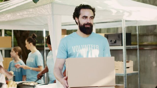 Kind young man donates non-perishable food items to the hungry and homeless as part of community food drive. Male caucasian volunteer carrying contribution boxes while looking at camera. Tripod shot.