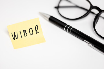 a yellow piece of paper stuck to a white table with the inscription "Wibor", next to it are glasses and a pen (selective focus)