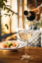 White wine poured into a glass with a served dish on the background at the restaurant 
