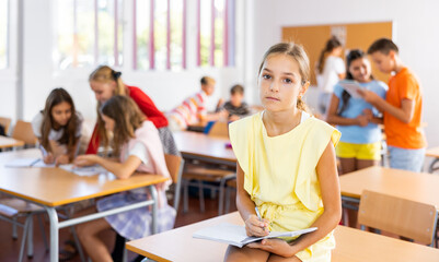 Upset tween girl sitting on table in schoolroom during recess on background with other pupils..