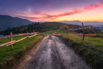 Fototapeta na wymiar Rural dirt road, wooden fence, green hills in mountain valley at sunset in autumn. Landscape with country road, meadows, trees, purple sky with pink clouds at twilight. Carpathians, Ukraine. Nature