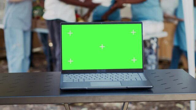Close-up of electronic laptop with isolated green screen sits on a table while charity workers assist the less privileged. Detailed view of minicomputer with a blank copyspace mockup template.