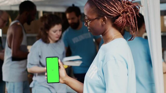Close-up of african american lady vertically grasping mobile device having blank copyspace chromakey template. During charitable food bank black woman volunteer holds cellphone with green screen.