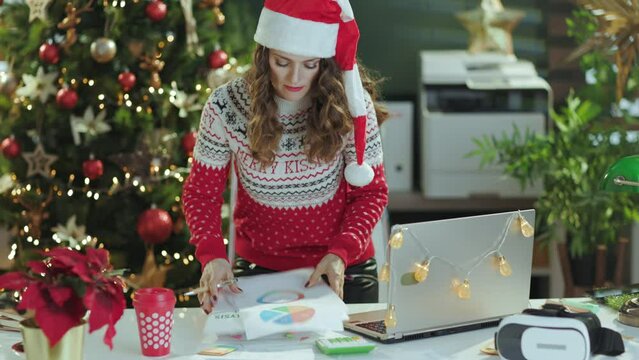 Christmas time. tired stylish middle aged small business owner woman in santa hat and red Christmas sweater with documents working in modern green office with Christmas tree.