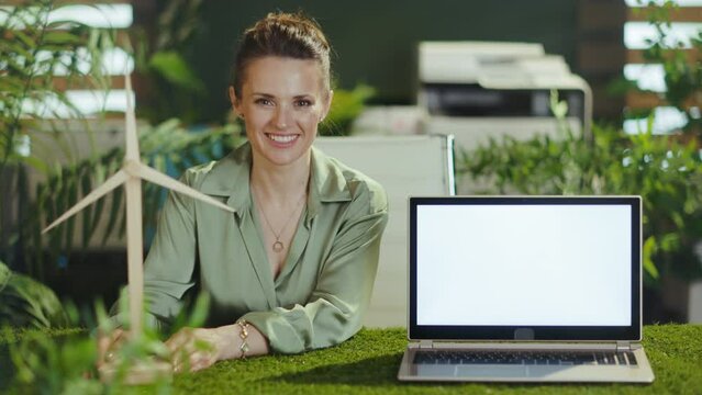 happy modern middle aged small business owner woman in green blouse in modern green office with windmill showing laptop blank screen.