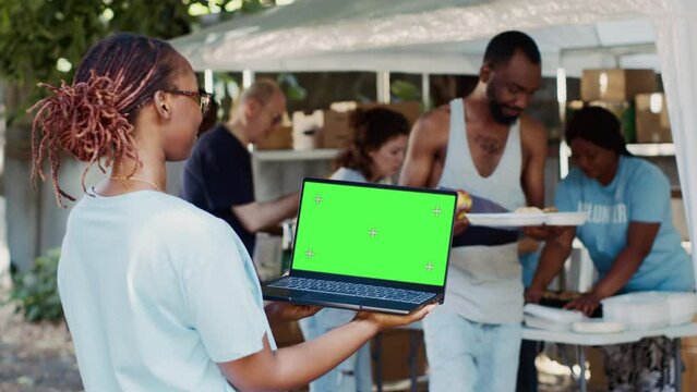 African american woman working for charity is shown holding laptop with blank chromakey template. Young black lady grasping wireless computer with isolated green screen for customization.