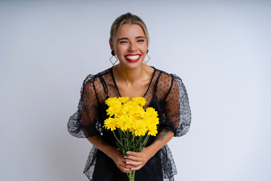 In an elegant black attire and captivating evening makeup, a stunning lady against white wall. Holding a bouquet of sunny yellow flowers, she radiates a festive and cheerful atmosphere
