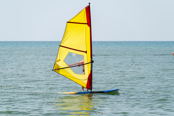 A man rides a blue board with a yellow sail in sunny weather at a tropical resort. A weak wind does not inflate the sail well.