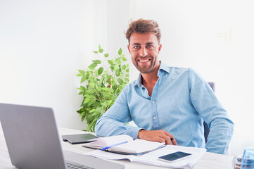 Positive attitude and motivation at work concept. Happy young professional man at his office desktop smiles looking at camera. - obrazy, fototapety, plakaty