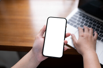A woman holding a smartphone while typing on keyboard, using a smartphone and laptop at her desk.