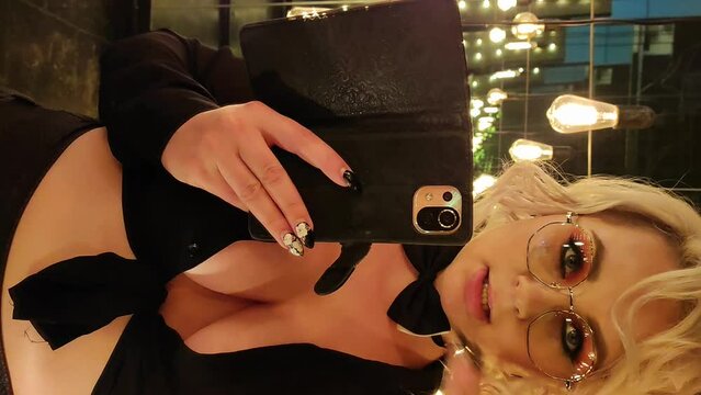 Portrait of sexy caucasian plus size blond woman wearing black blouse, skirt and bow tie and taking selfies in dark mirror hall with incandescent light bulbs decoration. Real time vertical video.