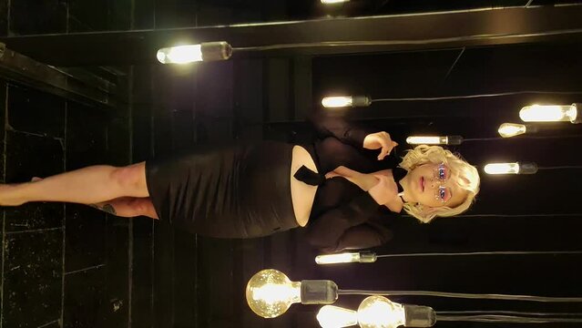Portrait of sexy caucasian plus size blond woman wearing black blouse, skirt and bow tie in dark mirror hall with incandescent light bulbs decoration. Real time handheld vertical video. Nightlife.