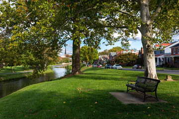 Tranquil scene in Frederick, MD: A park bench amid green grass by Carroll Creek offers a serene view of the city's quiet streets and distant church spires in the scenic urban landscape.