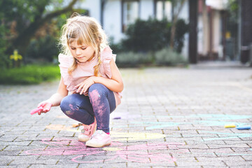 Little preschool girl painting with colorful chalks flowers on ground on backyard. Positive happy...
