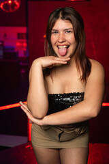 Fototapeta premium young caucasian woman posing from the front and showing her tongue on a background of red lights. night party concept.