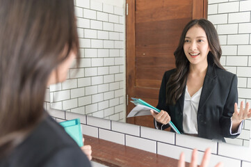 Young beautiful Asian woman in formal suit checking dressing tidiness and encourage herself in mirror at home before go for job interview. Woman recruitment employee or staff in company concept.