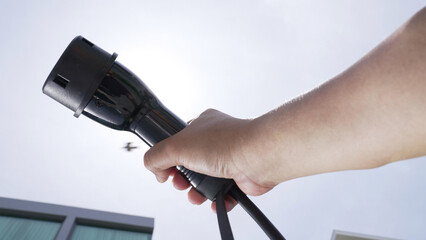Closeup hand grasping an EV plug for electric vehicle with the midday sky in the background as progressive idea of alternative sustainable clean and green energy for environmental concern.