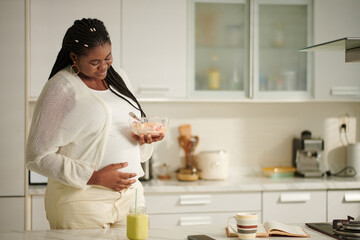 Smiling pregnant woman eating healthy breakfast and touching her belly