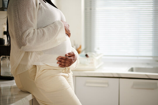 Cropped image of pregnant woman leaning on kitchen counter and touching her belly