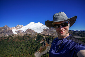 Adventurous athletic male hiker taking a selfie with the sun shinning and Mount Baker in the background.
