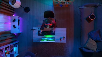 Top view of female in headphones sitting at the desk, hands on keyboard, streaming and playing game on computer online.