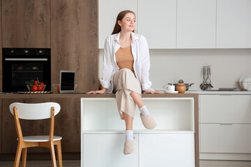 Beautiful young woman sitting on table with modern laptop in light kitchen