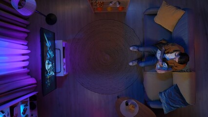 Top view of man sitting on the couch in the living room, watching movie on TV eating popcorn.