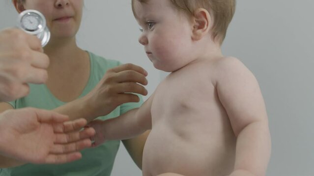 Mother puts baby on changing table and gives pacifier in mouth. Pediatrician uses stethoscope to listen to heartbeat and lungs of little child. Doctor does checkup in hospital. Close up. Slow motion.