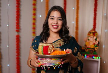 Obraz premium Beautiful Indian woman smiling at the camera on the occasion of Karwa Chauth - Indian rituals posing for camera. A young lady holding a decorated Karwa Chauth thali with a smile on her face - colo...