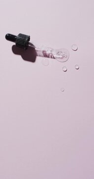 Vertical video of make up pipette and drops of serum with copy space on pink background