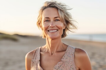Portrait of a smiling mature woman standing on the beach at sunset - obrazy, fototapety, plakaty