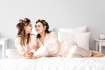 Little girl and her mother with hair curlers applying facial mask in bedroom